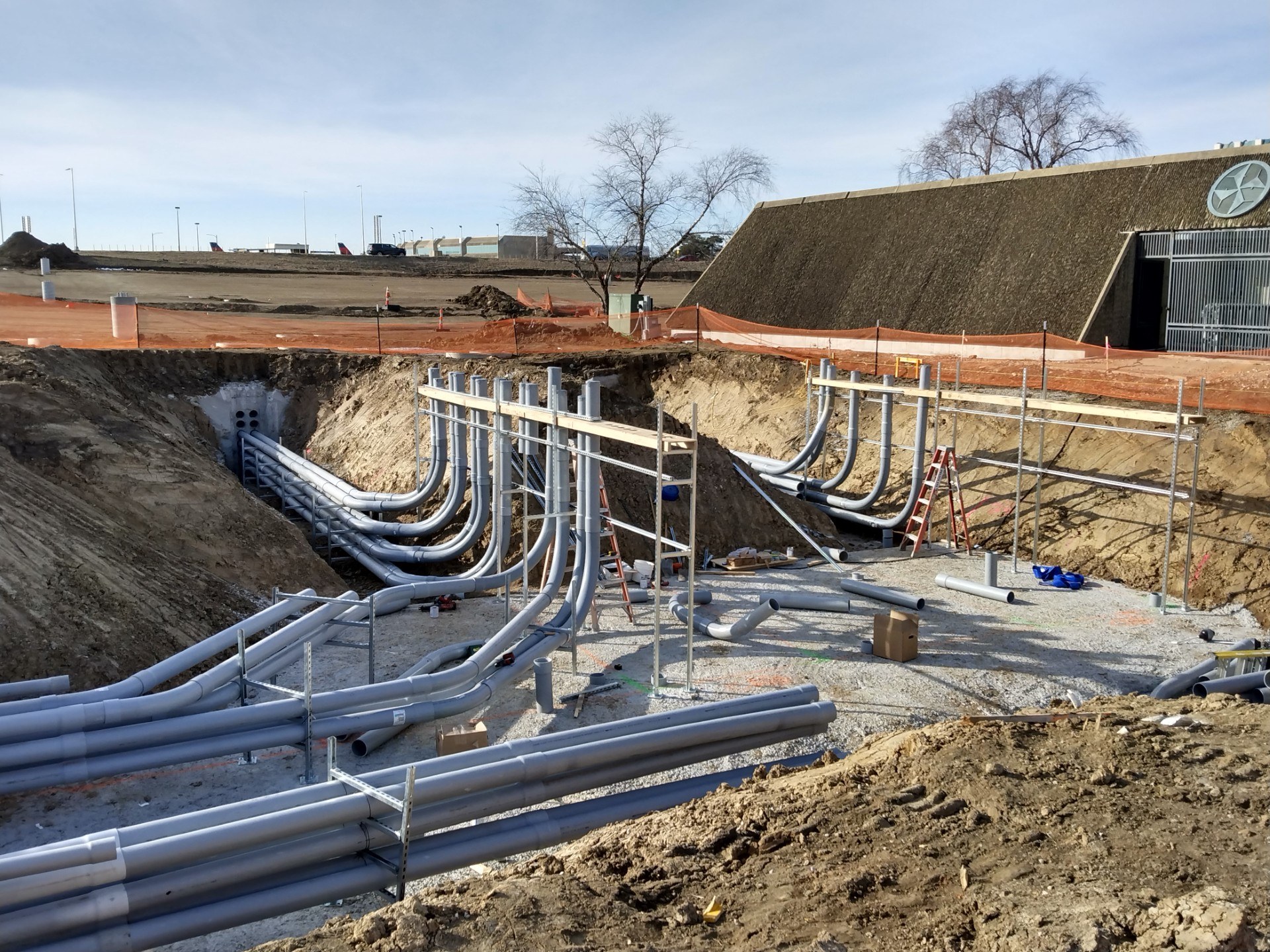 Racked PVC duct bank runs sweeping into an underground vault in an open excavation near an airport terminal