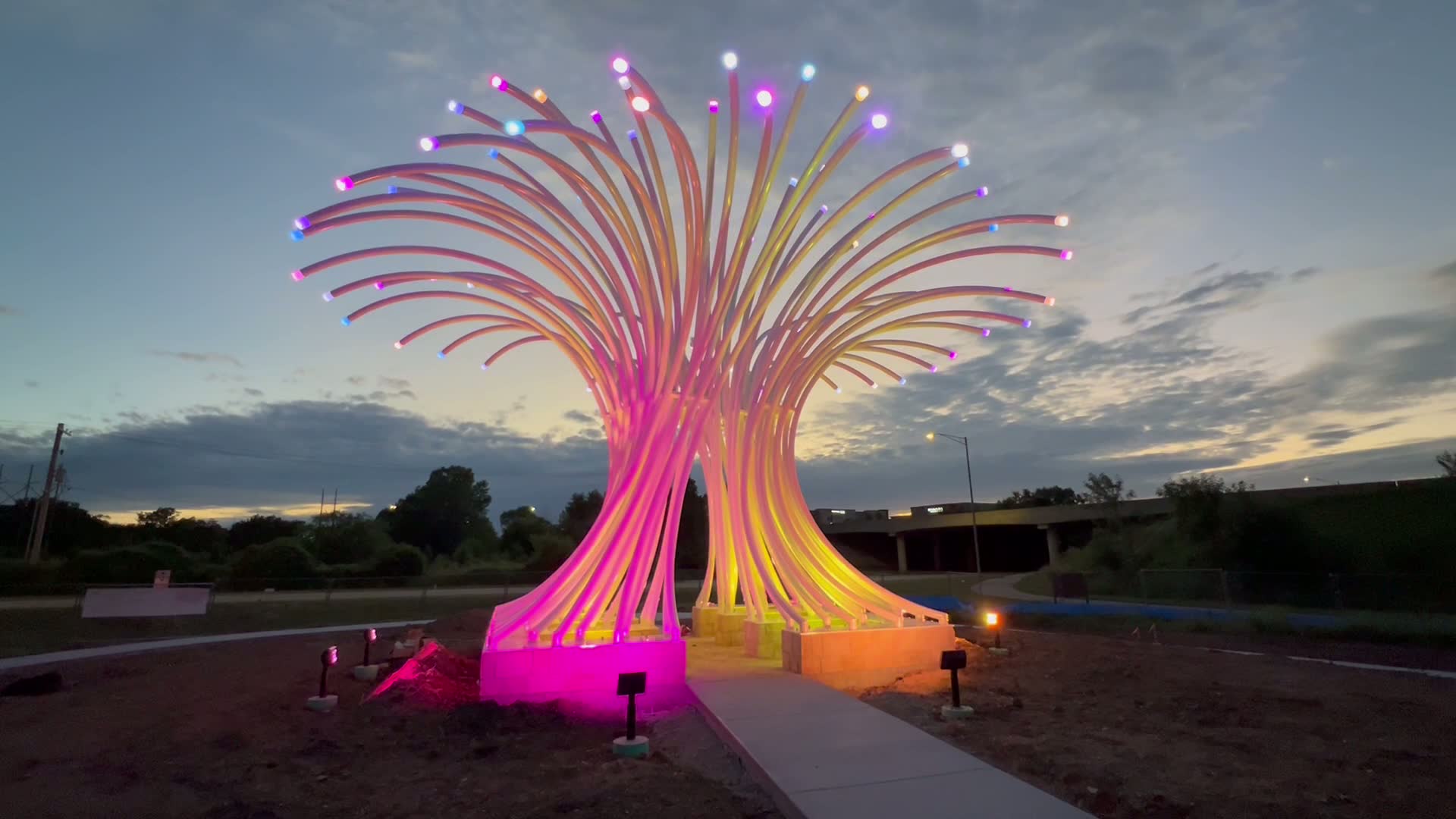 Large illuminated outdoor sculpture with color-changing LED arms at dusk